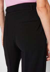 Personne portant un pantalon noir sur mesure avec une poche passepoilée à l'arrière et un haut violet clair rentré, montrée de la taille vers le bas.