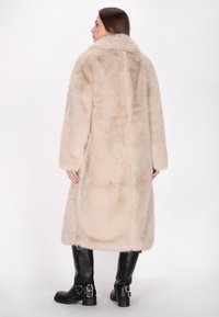 Lange, beige nepbontjas met een brede kraag en lange mouwen, met een zachte textuur en een rechte snit, gedragen over zwarte kniehoog laarzen.