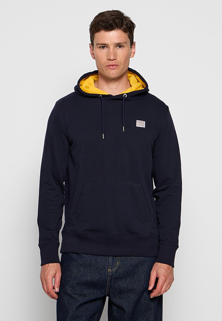 Homme portant un hoodie bleu marine avec une doublure jaune à capuche et un petit patch sur la poitrine, associé à un jean bleu foncé, se tenant devant un fond uni.