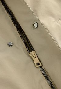 Beige jakke stof med en synlig sort lynlås, en crème lynlåsgreb og sølvnapsknapper nær åbningen. Glat tekstur, minimalistisk design.