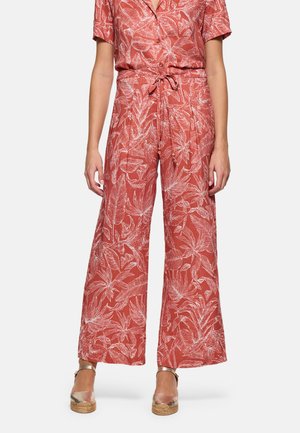 Mujer con pantalones de pierna ancha y tiro alto en color óxido con un estampado floral blanco, cintura atada, combinados con sandalias de cuña doradas.