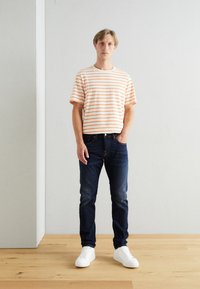 T-shirt a righe con collo a girocollo in crema e strisce arancioni, indossata con jeans blu scuro. Il modello indossa sneaker bianche, in piedi su un pavimento di legno.