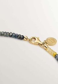 Cierre de langosta dorado sujeto a una pulsera de cuentas con piedras facetadas azules y amarillas y una pequeña etiqueta redonda dorada.