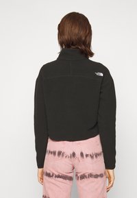 Persona con capelli corti castani indossa una giacca in pile nera della North Face e jeans rosa tie-dye, in piedi con le spalle rivolte verso la fotocamera su uno sfondo semplice.
