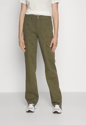 Persoon die olijfgroene wijde cargobroek en witte sneakers draagt, staand tegen een eenvoudige lichte achtergrond.