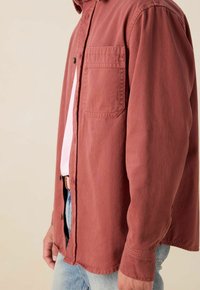 Roestbruine button-up shirt van lichtgewicht katoen met een borstzak, gebogen zoom en lange mouwen, gedragen over een roze t-shirt.