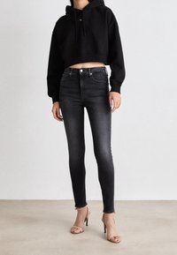 Sudadera recortada negra con cordón, combinada con jeans ajustados de talle alto en gris oscuro y tacones nude con tiras. Texturas suaves en todo.