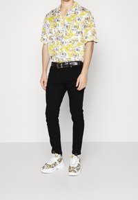 Kortärmad vit skjorta med grafisk textdesign i gult och svart, tillsammans med svarta skinny jeans och mönstrade vita sneakers.