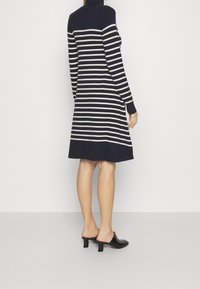 Nainen, jolla on yllään tummansiniraidallinen pitkähihaisen poolopaidan mekko ja mustat korkeat mules-kengät, seisoo vaaleaa taustaa vasten.