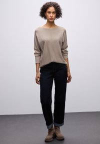Maglione beige chiaro a maglia larga con scollo a barca, polsini e cucitura centrale; abbinato a jeans a gamba dritta blu scuro e stivaletti marroni.