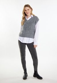 Grauer gestrickter ärmelloser Pullover mit V-Ausschnitt, über einem weißen Hemd getragen, kombiniert mit dunkelgrauen Hosen und schwarzen Stiefeletten.