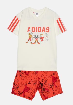T-shirt crema con testo rosso "ADIDAS" e grafica di un personaggio; pantaloni corti rossi con stampa di personaggi dei cartoni animati. Mix di cotone, maniche corte e scollo a v.