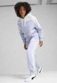 Tuta di colore viola chiaro con cappuccio bianco, caratterizzata da un logo testurizzato sul petto. I pantaloni hanno strisce laterali e sono abbinati a sneakers bianche.