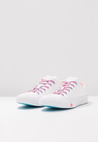 Białe płócienne trampki z różowymi i niebieskimi sznurówkami, okrągły czubek, niskoprofilowy design oraz różowa gwiazda na krawędzi zewnętrznej podeszwy.