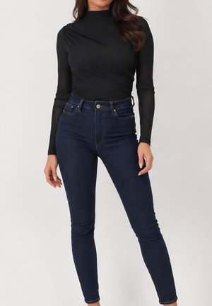 Vrouw draagt een zwarte aansluitende top met lange mouwen en donkerblauwe high-waisted skinny jeans tegen een effen achtergrond.