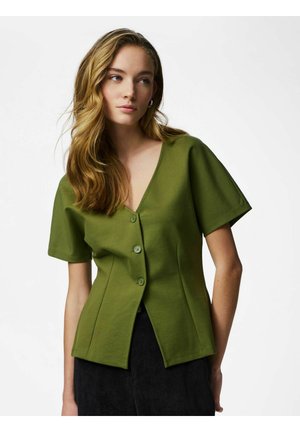 Jeune femme aux longs cheveux ondulés portant un chemisier vert à manches courtes boutonné et un pantalon noir sur un fond uni.