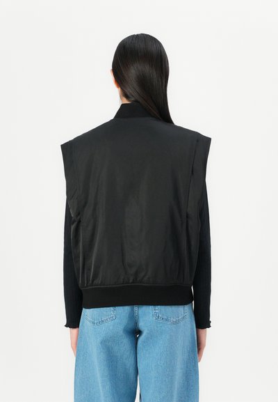 Giacca nera senza maniche con una texture liscia, colletto a coste e orlo elastico. Indossata sopra un top nero a maniche lunghe e jeans blu.