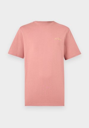 Lihtne tuhm roosa lühikeste varrukatega T-särk väikese kollase kursiivkirjas "Les Deux" logoga ülemisel vasakul rinnal valgel taustal.