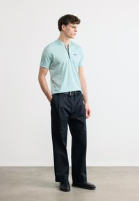 Νεαρός άνδρας που στέκεται, φοράει ανοιχτό μπλε μπλουζάκι με φερμουάρ και κοντά μανίκια, μαύρη ζώνη, σκούρο φαρδύ παντελόνι και μαύρα παπούτσια, μπροστά σε απλό φόντο.