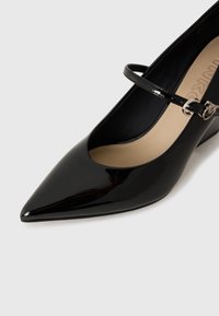 Escarpin en cuir verni noir avec un bout pointu, une lanière de cheville avec une boucle métallique et une semelle intérieure beige. Texture lisse.