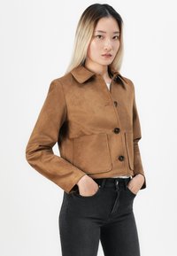 Giacca cropped in finta suede marrone con colletto, dotata di due tasche frontali e bottoni neri. Abbinata a jeans neri a vita alta.