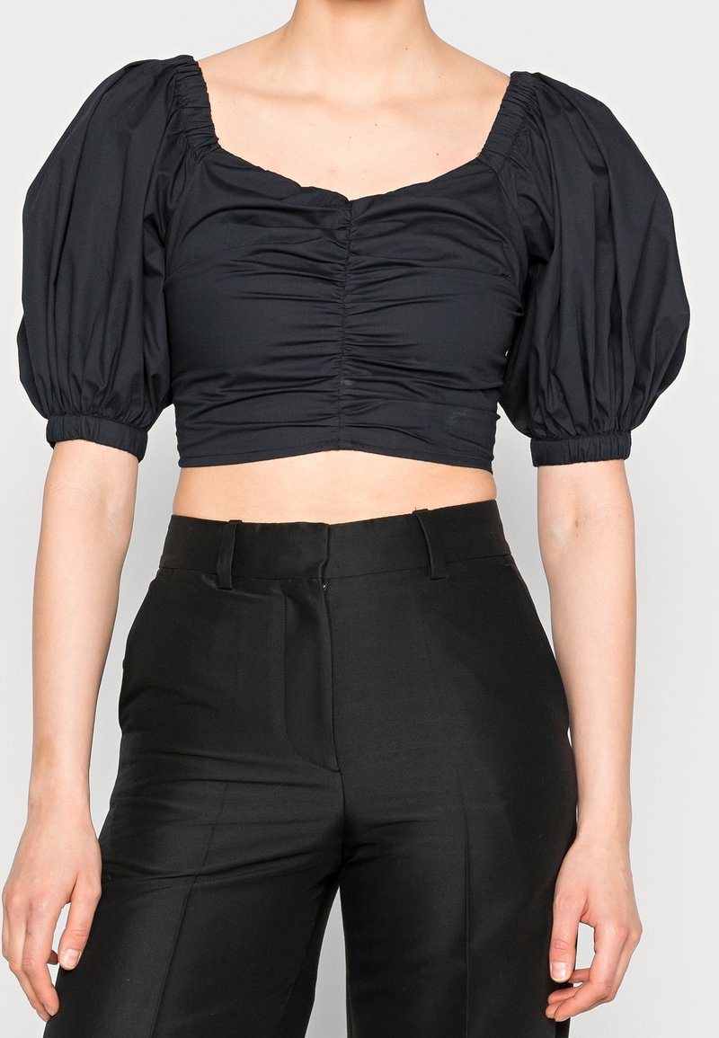 Blusa negra cortada con mangas abullonadas, que presenta un escote cuadrado y textura fruncida en la parte delantera. Combinada con pantalones negros de cintura alta.