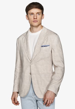 Giovane uomo con un blazer beige sopra una camicia bianca e jeans azzurri chiari, in piedi e che guarda leggermente di lato su uno sfondo semplice.