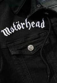 Svart denimjacka med vitt broderat "Motörhead"-logotyp, silverfärgad dödskalleknapp på bröstfickan, texturerat tyg och dragsko.
