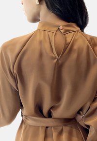 Abito in satin marrone con schiena a occhiello, vita con cintura e dettagli arricciati. Presenta un piccolo bottone decorativo sul colletto.