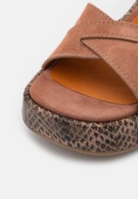 Sandale en daim marron avec un design de bride courbée et une semelle texturée au motif reptilien foncé. L'intérieur présente une semelle intérieure orange lisse.