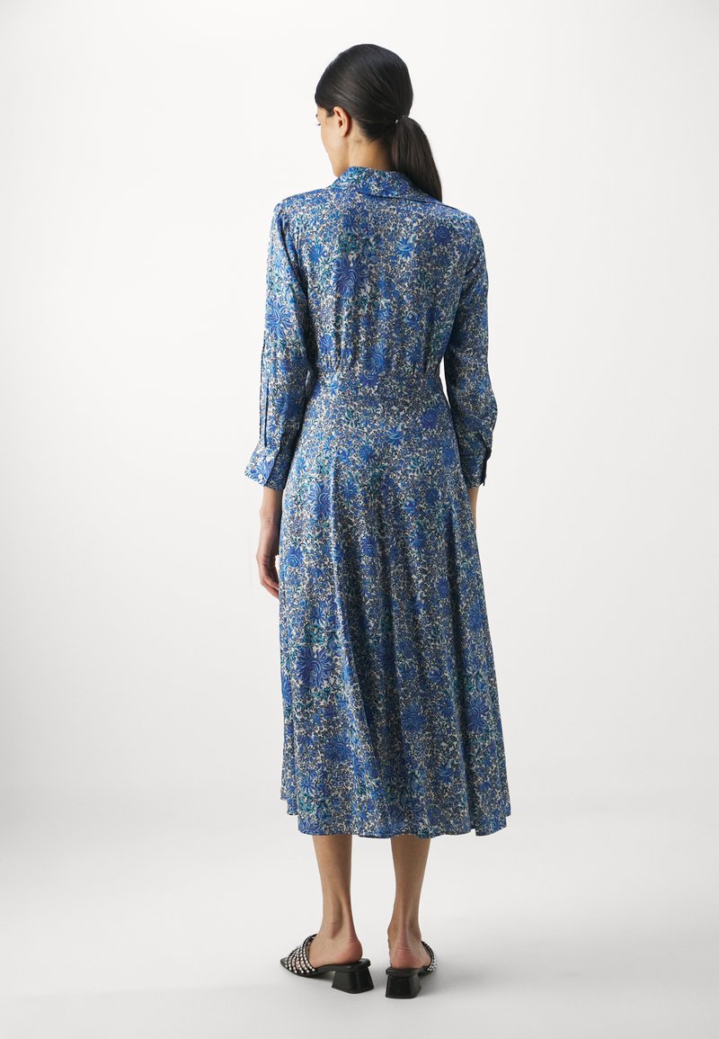 Midikleid im blauen Blumenmuster, aus leichtem Stoff gefertigt. Merkmale sind lange Ärmel, taillierter Schnitt und ausgestellter Rock. Schwarze High Heels.