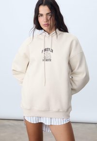 Vaučasti oversized dukserica s kapuljačom na vezanje, s grafikom "FOREVER MY BESTIE" i dva crtana životinja na prednjoj strani.