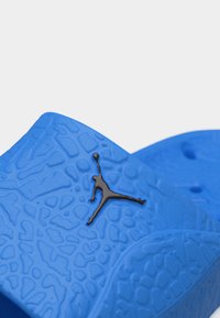 Μπλε σατινέ σαγιονάρα με μαύρο λογότυπο Jumpman στο λουράκι, κοντινή όψη σε ανοιχτό φόντο.