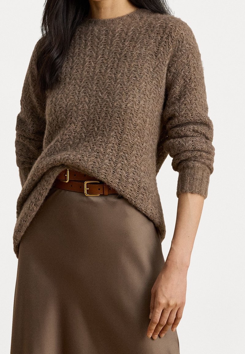 Femme portant un pull en maille marron texturé, rentré dans une jupe marron soyeuse avec une ceinture beige à boucle dorée.
