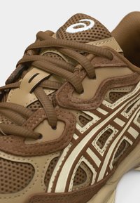 Baskets marron et beige en mesh et suède avec des lacets épais, un panneau latéral à motifs et un logo blanc sur la languette rembourrée.