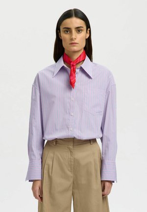 Kvinde med glat mørkt hår iført en pastelstribet skjorte med knapper, beige bukser med høj talje og et rødt tørklæde bundet om halsen, vendt fremad.