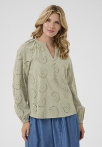 Hellgrüne Bluse mit gesticktem Paisley-Muster, Knopfleiste und elastischen Bündchen. Kombiniert mit einem Jeansrock.