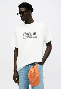 Άνδρας με μαύρα γυαλιά, λευκό μπλουζάκι με το λογότυπο "Carhartt work in progress", μπλε τζιν, με πορτοκαλί καπέλο που κρέμεται από την τσέπη του, σε λευκό φόντο.