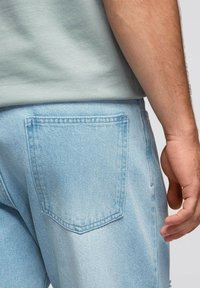 Jeans de denim azul claro con un bolsillo trasero rectangular, detalles sutiles de costura y un ajuste relajado. Llevados con una camisa verde.