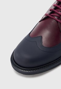 Bőr cipő kéttónusú kialakítással; bordó és tengerészkék színekben. Sima textúrával, hullámos mintázattal és bordó fűzővel rendelkezik.