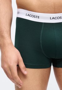 Άνδρας που φοράει σκούρα πράσινα αντρικά εσώρουχα Lacoste με λευκή ελαστική ζώνη μέσης που αναγράφει το όνομα της μάρκας, με το χέρι ακουμπισμένο δίπλα στον μηρό.