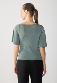 Top cu mânecă scurtă, cu o croială lejeră, într-o nuanță de verde închis, având o textură netedă și decolteu larg, asociat cu pantaloni negri.
