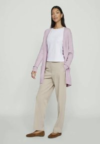 Cárdigan de color púrpura claro llevado sobre una camiseta blanca, combinado con pantalones beige y zapatos marrones sin cordones. Tela suave con diseño de apertura frontal.