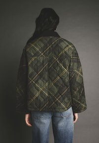 Jachetă matlasată de culoare verde închis, cu un model carat galben, având un guler ribbit și o croială lejeră, asociată cu blugi din denim albastru.