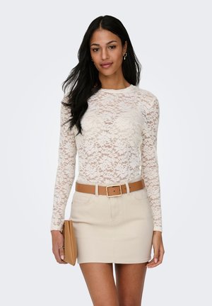 Donna che indossa un top in pizzo crema a maniche lunghe, mini gonna beige con cintura color cuoio, che tiene in mano una pochette color cuoio, in piedi davanti a uno sfondo chiaro e uniforme.