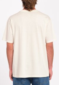 Camiseta de manga corta en color crema con un corte relajado, que presenta un diseño liso en la parte posterior y está confeccionada con una tela suave y ligera. Se lleva con unos vaqueros azules.