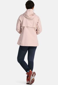Vaaleanpunainen vedenpitävä takki hupulla ja takana läppäyksellä, yhdistettynä tummiin legginsseihin ja punaisiin vaelluskengiin.