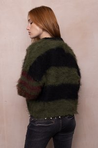 Junge Frau mit langen roten Haaren, die einen flauschigen, gestreiften grünen und schwarzen Pullover, dunkle Jeans und goldene Ohrringe trägt und seitlich vor einer schlichten Wand steht.