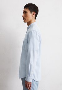 Camicia azzurra a righe con colletto button-down, maniche lunghe, taschino sul petto e tessuto di cotone liscio, indossata da una persona di profilo.