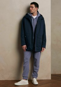 Granatowa kurtka z kapturem, z dwoma kieszeniami z przodu, nałożona na jasnoszary hoodie i pasujące dresy. Białe sneakersy dopełniają całość.